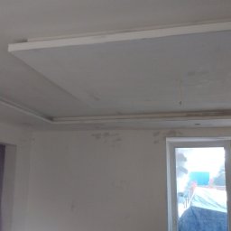 Usługi ogólnobudowlane - Wykończony sufit podwieszany z listwami dekoracyjnymi w odświeżonym pokoju. Widoczny grzejnik i okno. Ściany przygotowane do malowania.