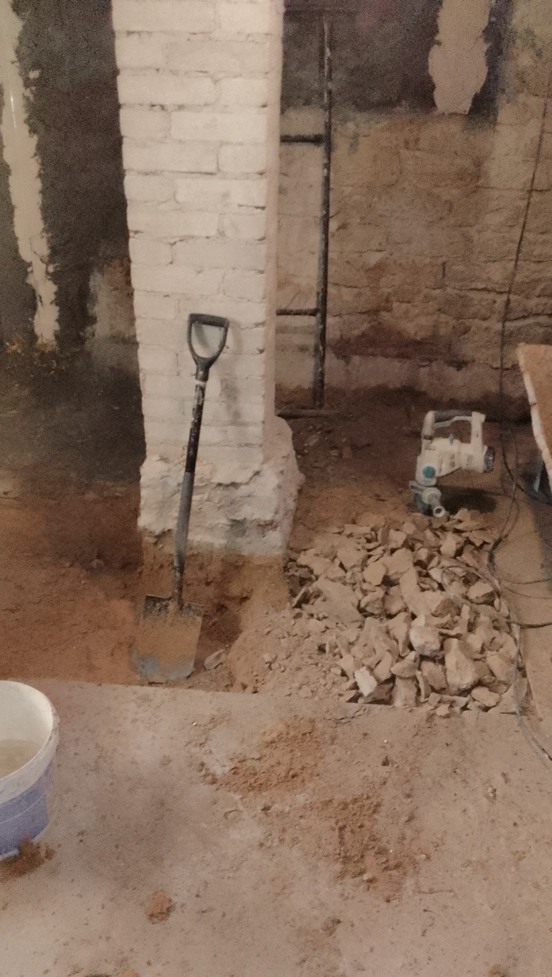 Etap prac remontowych: wykopana podłoga przy ceglanym filarze, łopata wbita w ziemię, gruz i młot pneumatyczny w tle. Przygotowanie do wylewki.