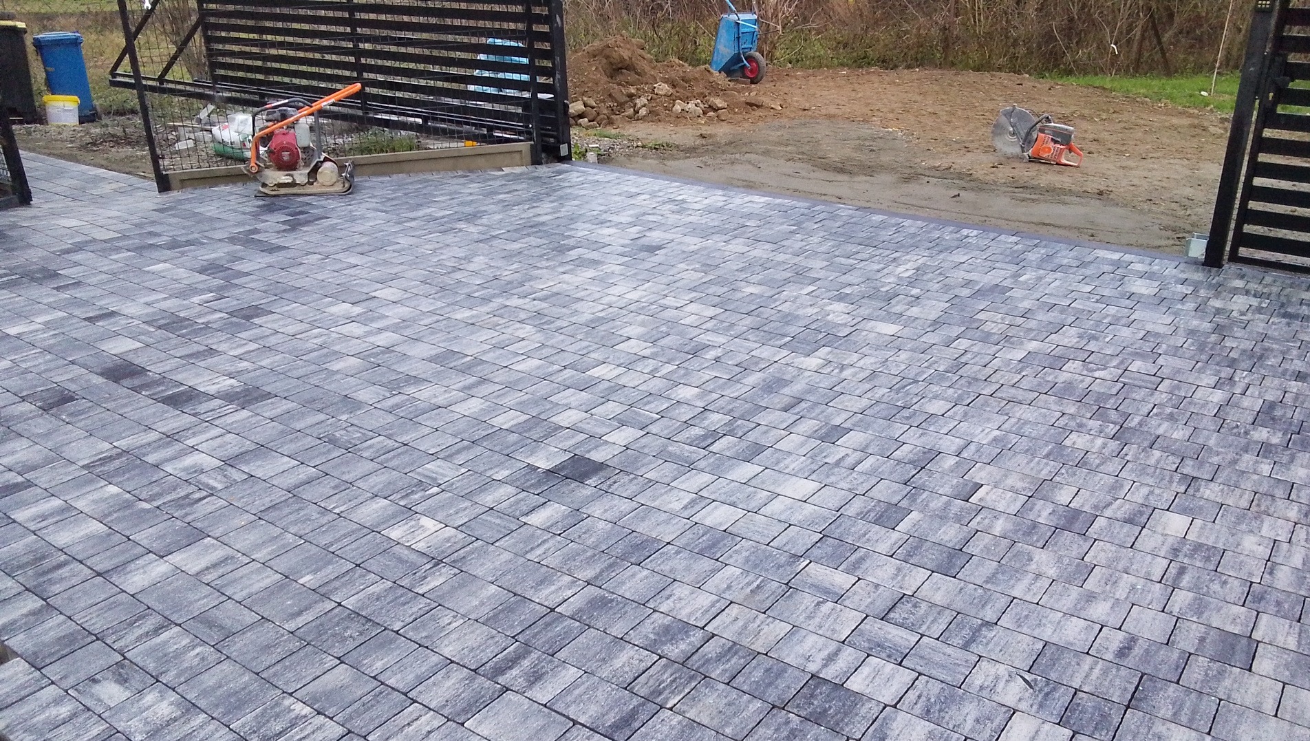 Ułożona szara kostka brukowa na podjeździe, widoczna zagęszczarka, piła do cięcia kostki i taczka w tle, brama wjazdowa.