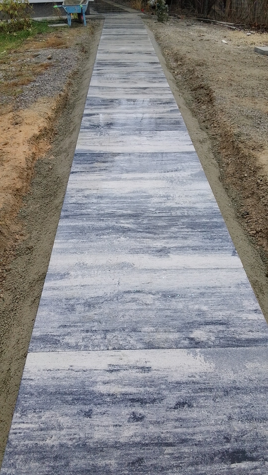 Ułożony chodnik z szarych płyt imitujących drewno. Widoczna taczka w tle. Świeżo wykonana ścieżka w ogrodzie.