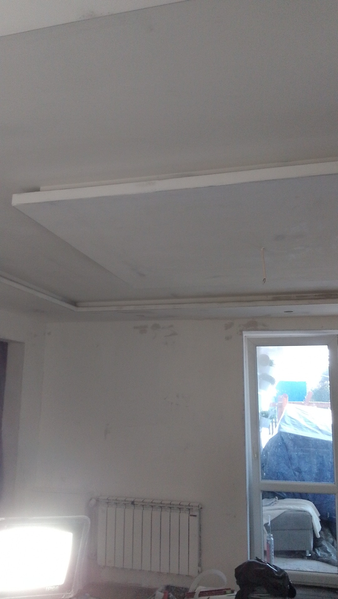 Wykończony sufit podwieszany z listwami dekoracyjnymi w odświeżonym pokoju. Widoczny grzejnik i okno. Ściany przygotowane do malowania.