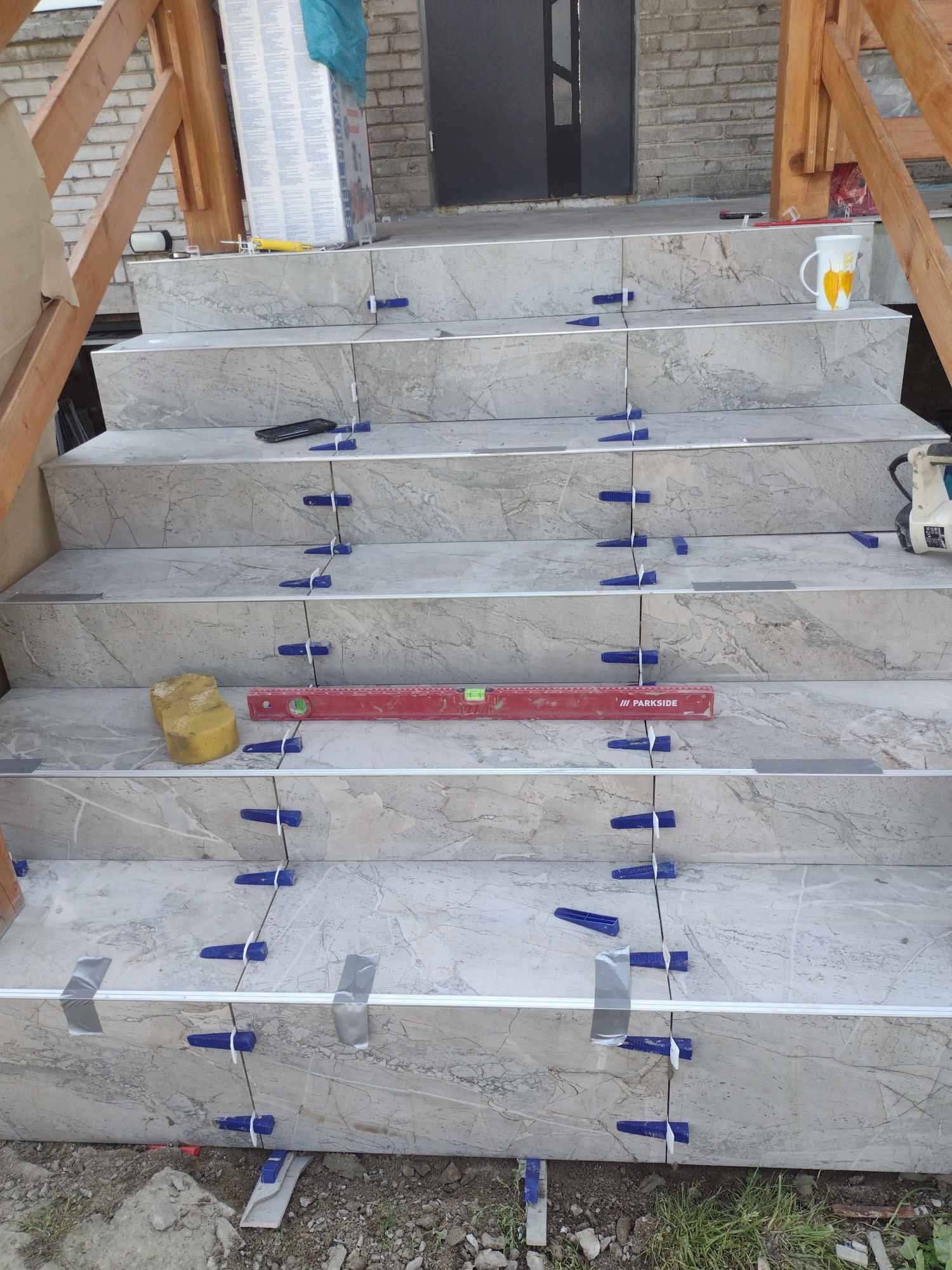 Wykończenie schodów wejściowych płytkami imitującymi marmur. Widoczne poziomica, gąbka, kliny do płytek i listwy. Etap prac przed fugowaniem.