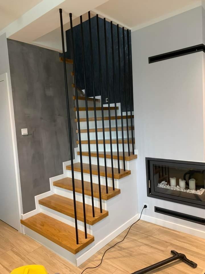 Nowoczesne schody drewniane z czarną balustradą w jasnym wnętrzu. Widoczny kominek z dekoracją. Podłoga drewniana. Ściana szara z efektem betonu.