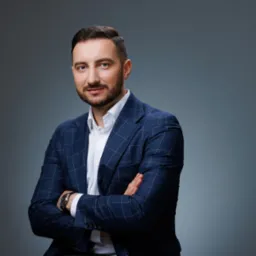 Mężczyzna w niebieskiej marynarce w kratę, biała koszula, zegarek na ręku, skrzyżowane ramiona, na szarym tle, patrzy wprost, profesjonalny portret.