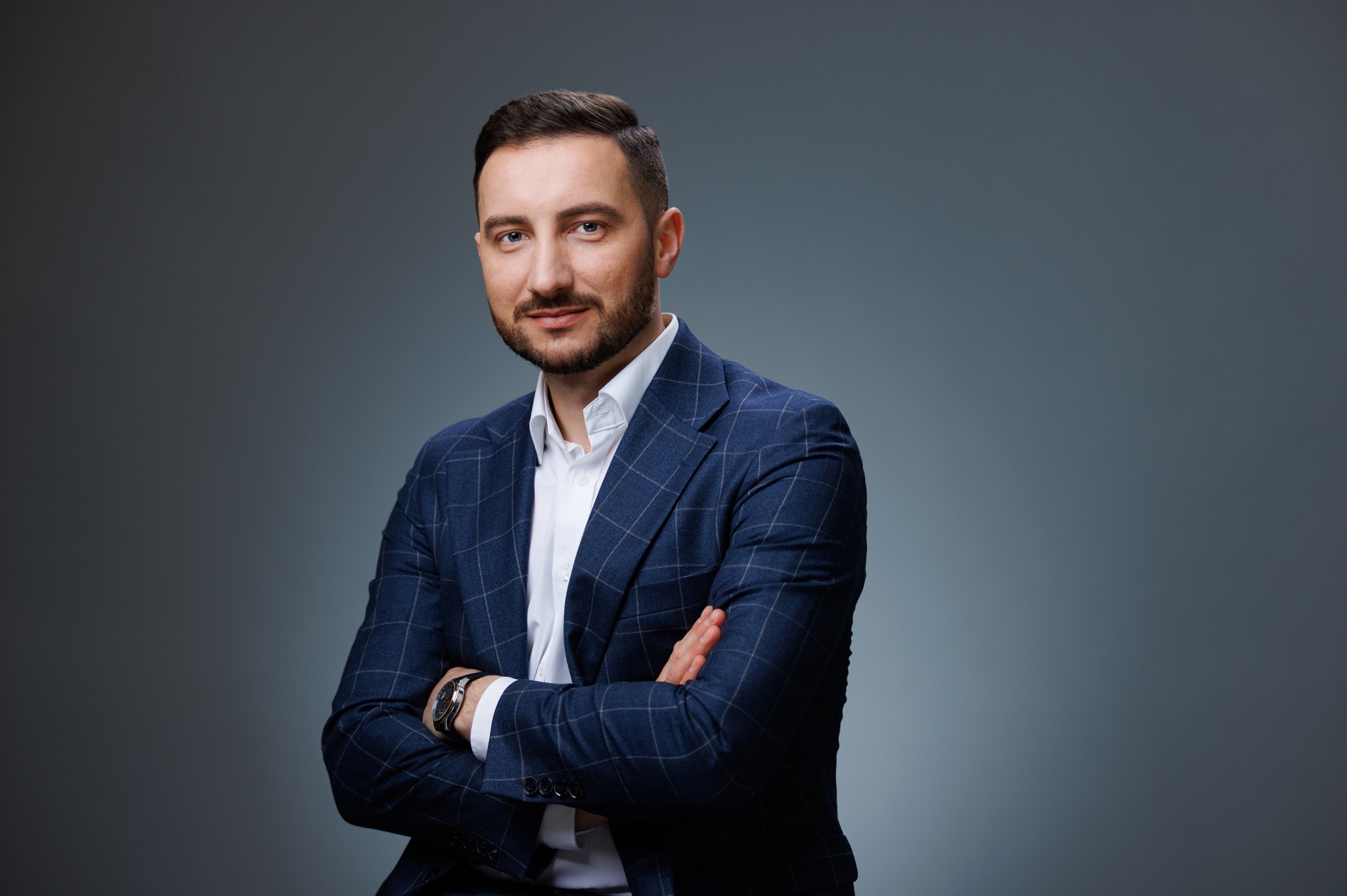 Mężczyzna w niebieskiej marynarce w kratę, biała koszula, zegarek na ręku, skrzyżowane ramiona, na szarym tle, patrzy wprost, profesjonalny portret.