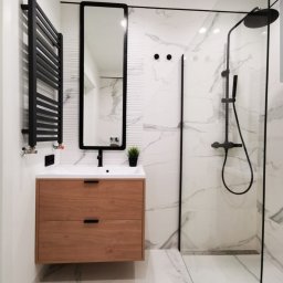 Andrzej Miętki - Nowoczesna łazienka z drewnianą szafką pod umywalką, czarnym grzejnikiem drabinkowym i kabiną prysznicową typu walk-in z armaturą w czarnym macie.