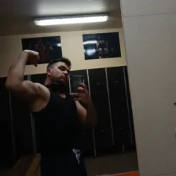 Mężczyzna w czarnym tank topie pozuje do selfie w lustrze siłowni, prezentując biceps. W tle szafki i plakaty z motywami fitness.