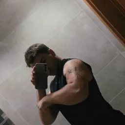 Mężczyzna w czarnej koszulce robi selfie, eksponując wytatuowany biceps. Widoczna część łazienki z szarą glazurą i fragmentem szafki.