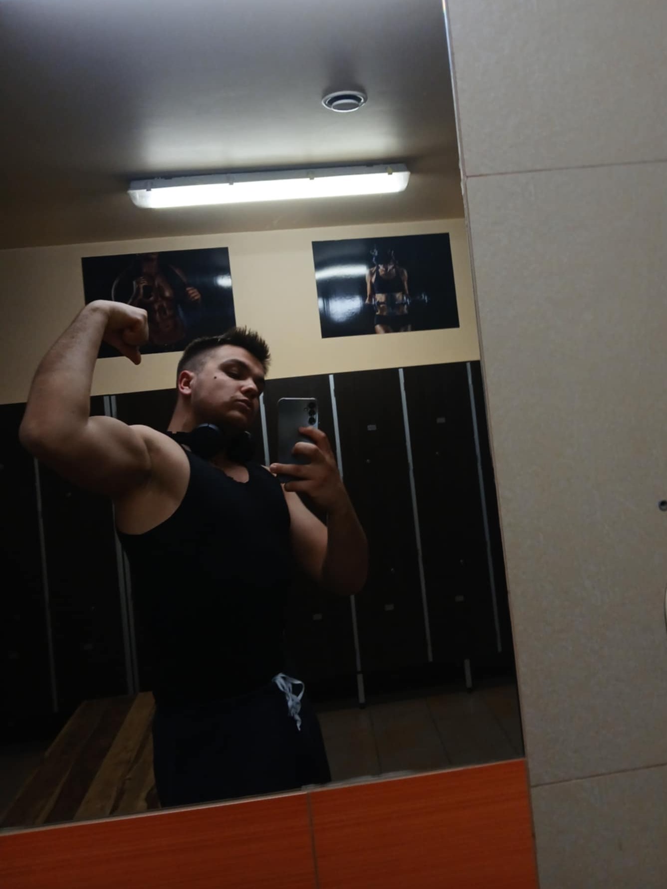 Mężczyzna w czarnym tank topie pozuje do selfie w lustrze siłowni, prezentując biceps. W tle szafki i plakaty z motywami fitness.