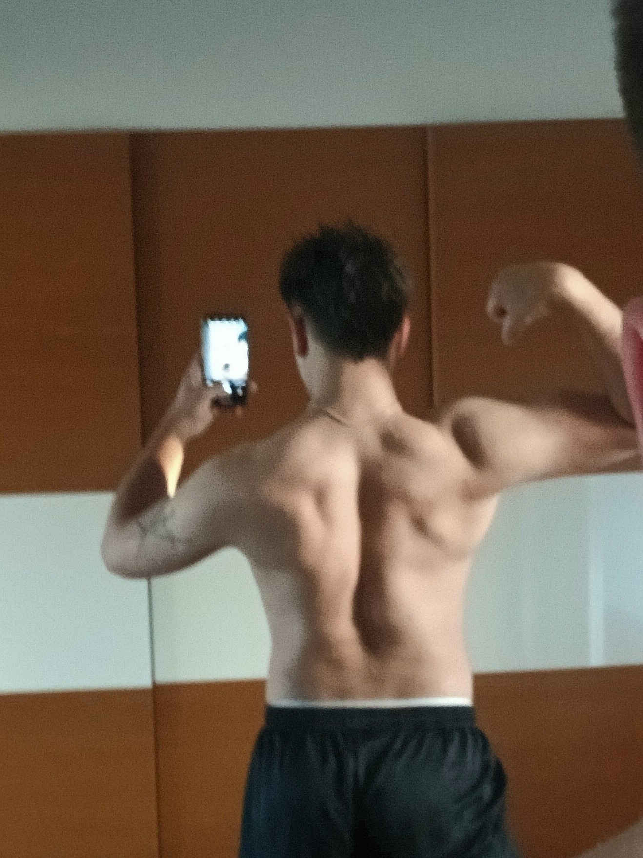 Mężczyzna z widocznymi mięśniami pleców, pozujący do zdjęcia telefonem. Widoczny tatuaż na ramieniu, czarne spodenki i fragment osoby po prawej stronie kadru.