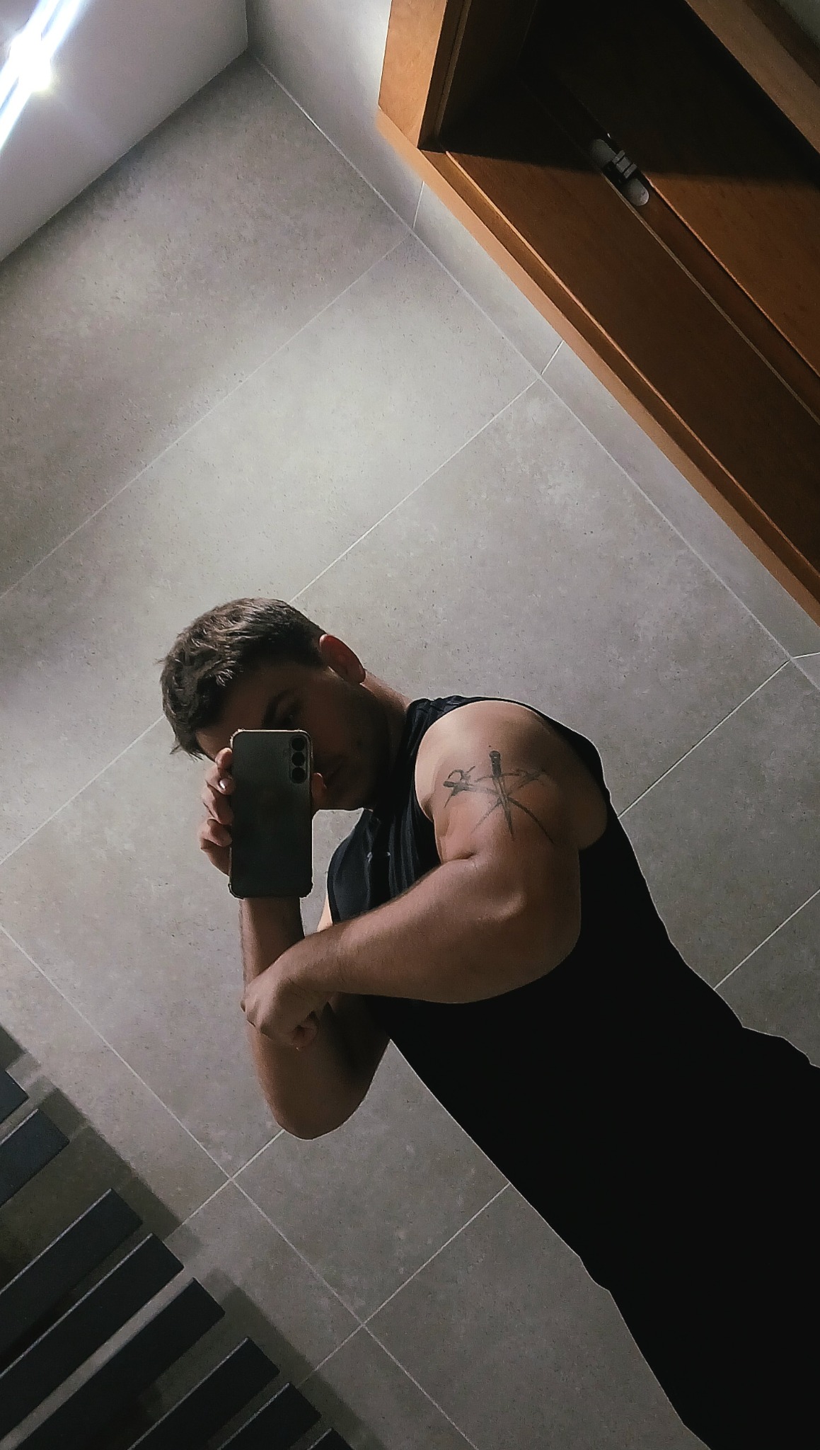 Mężczyzna w czarnej koszulce robi selfie, eksponując wytatuowany biceps. Widoczna część łazienki z szarą glazurą i fragmentem szafki.