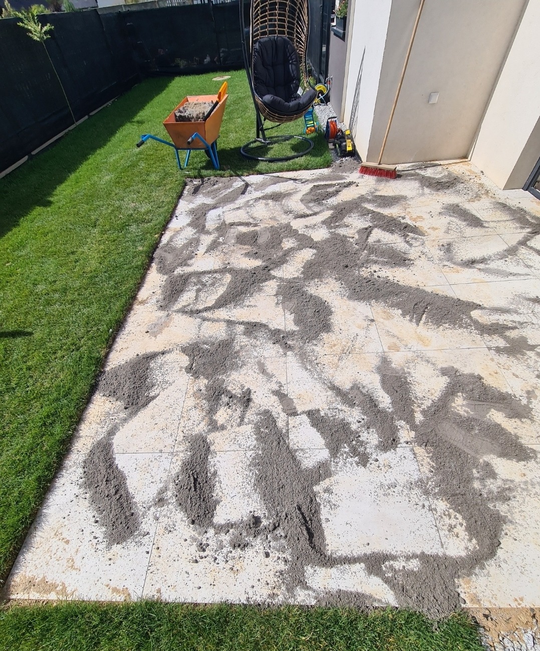 Taras w trakcie budowy: jasne płytki z rozsypanym piaskiem, obok trawnik, taczka z narzędziami i wiszący fotel. Widoczne narzędzia do układania nawierzchni.