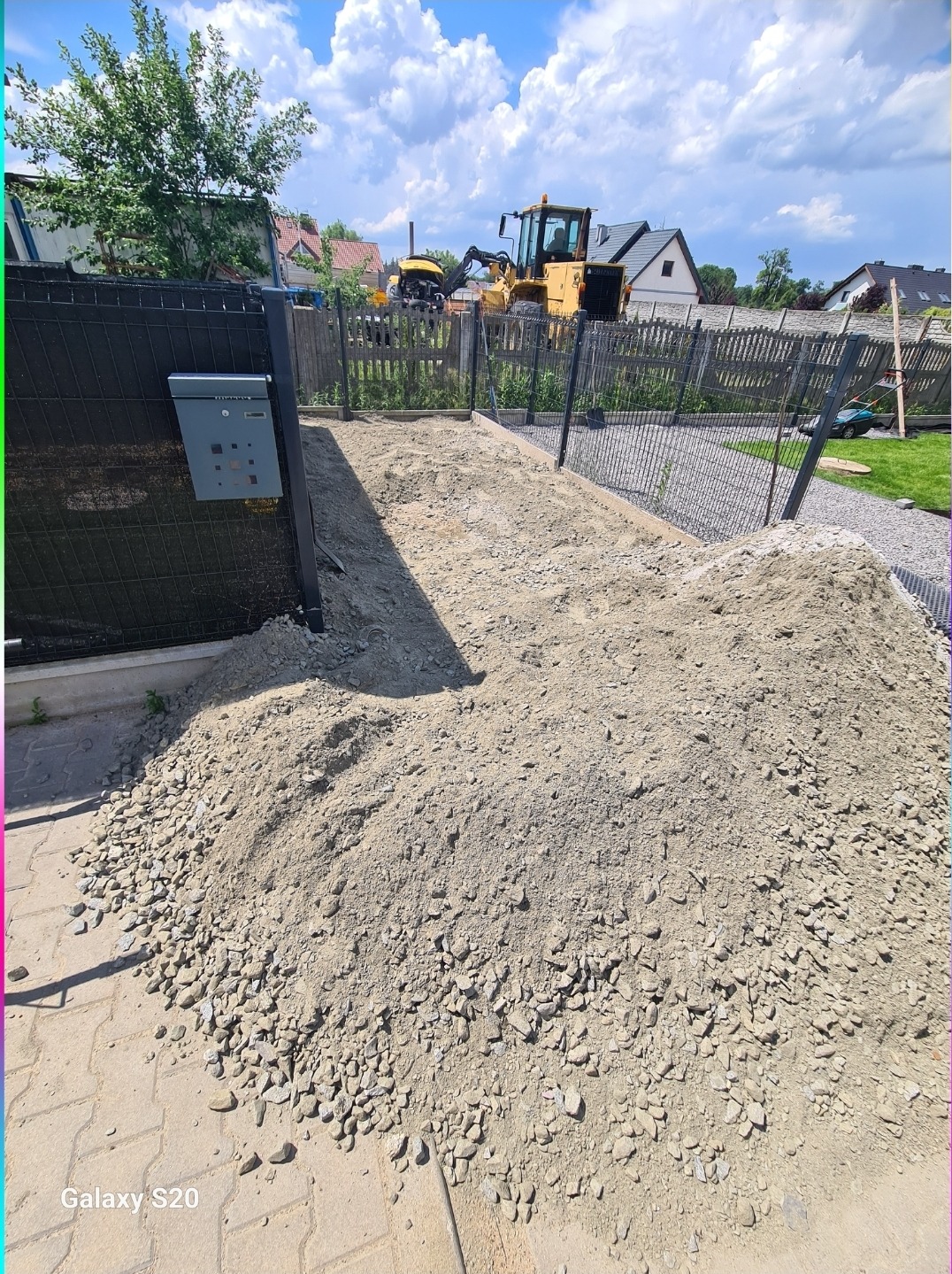 Przygotowanie podłoża pod podjazd: wysypany żwir, w tle maszyny budowlane i ogrodzenie. Widok z bliska na etap prac ziemnych.