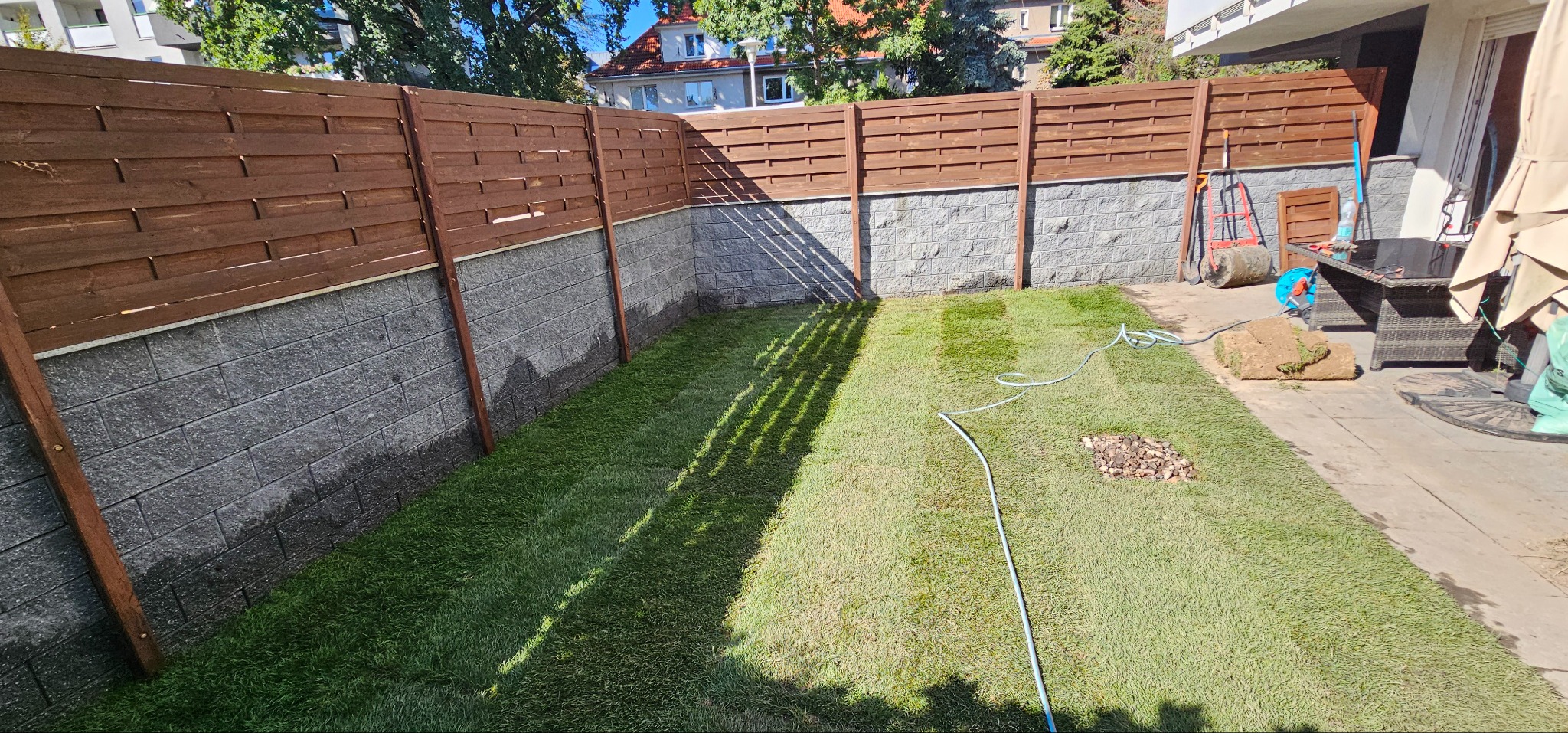Fragment ogrodu po założeniu trawnika z rolki, widoczny drewniany płot z podmurówką z szarej kostki, narzędzia ogrodnicze i zwinięte rolki darni w tle.