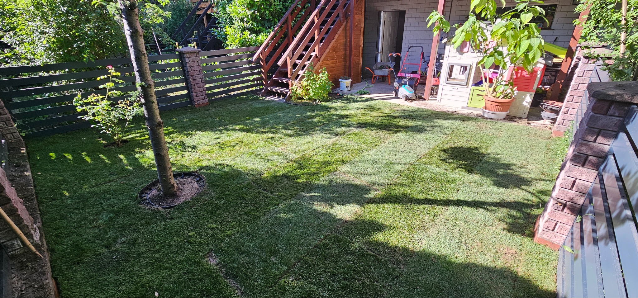 Świeżo ułożona, soczysta zieleń trawy w rolkach w zacisznym ogrodzie, z widocznym drzewem i elementami architektury ogrodowej w tle. Cień drzewa dodaje głębi.