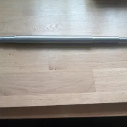 Metalowy wał z gwintowanymi końcami leżący na jasnym drewnianym blacie. Widoczne ślady obróbki. Detal toczony z precyzyjnie wykonanym gwintem.