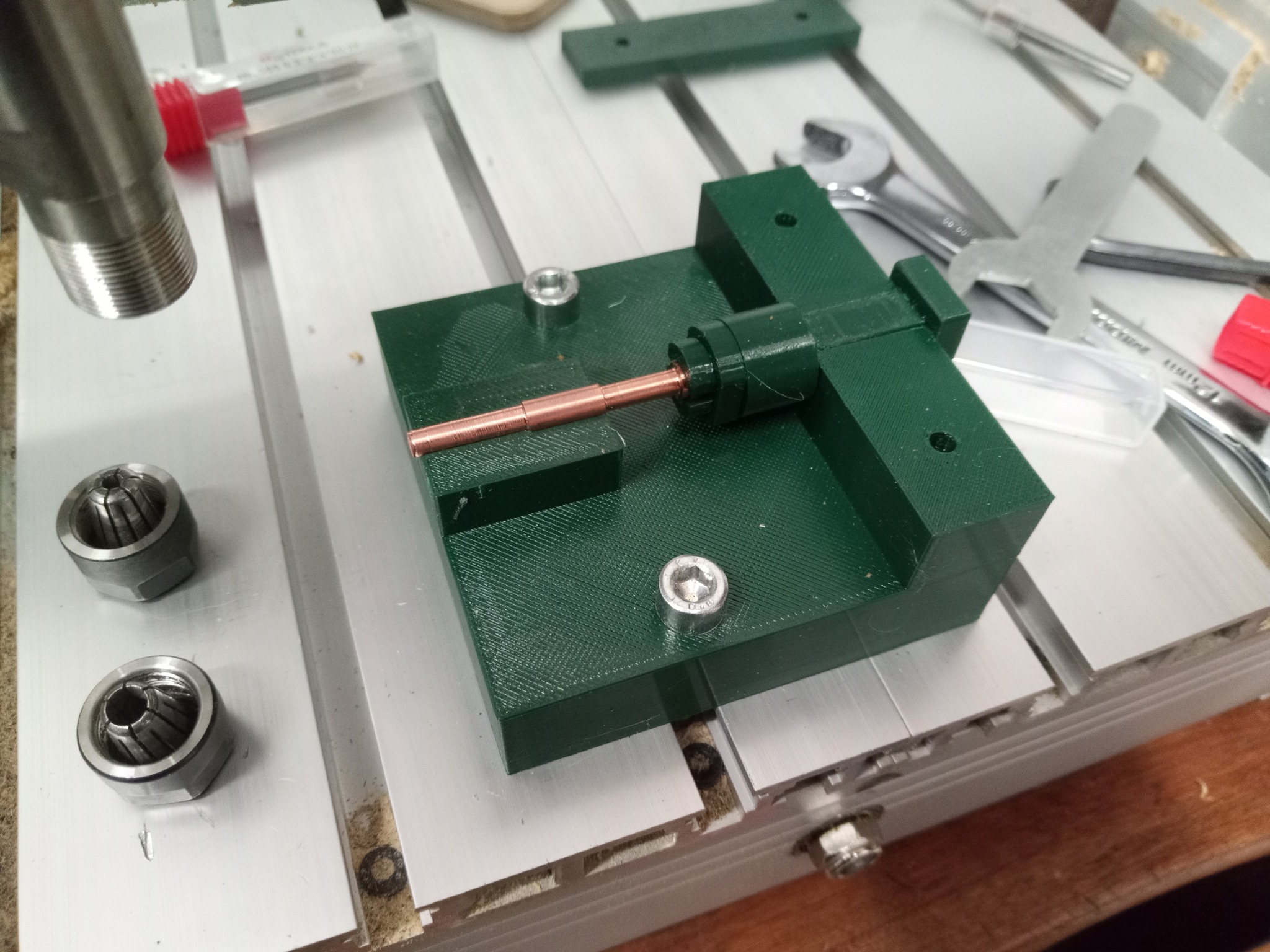 Zielony uchwyt z mosiężnym prętem na aluminiowym stole warsztatowym, obok klucze i tuleje zaciskowe. Detal z bliska, oświetlenie naturalne.