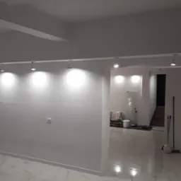 Białe, świeżo pomalowane wnętrze z marmurową podłogą i oświetleniem punktowym. Widoczne worki z materiałami budowlanymi w tle, sugerujące etap wykończeniowy.