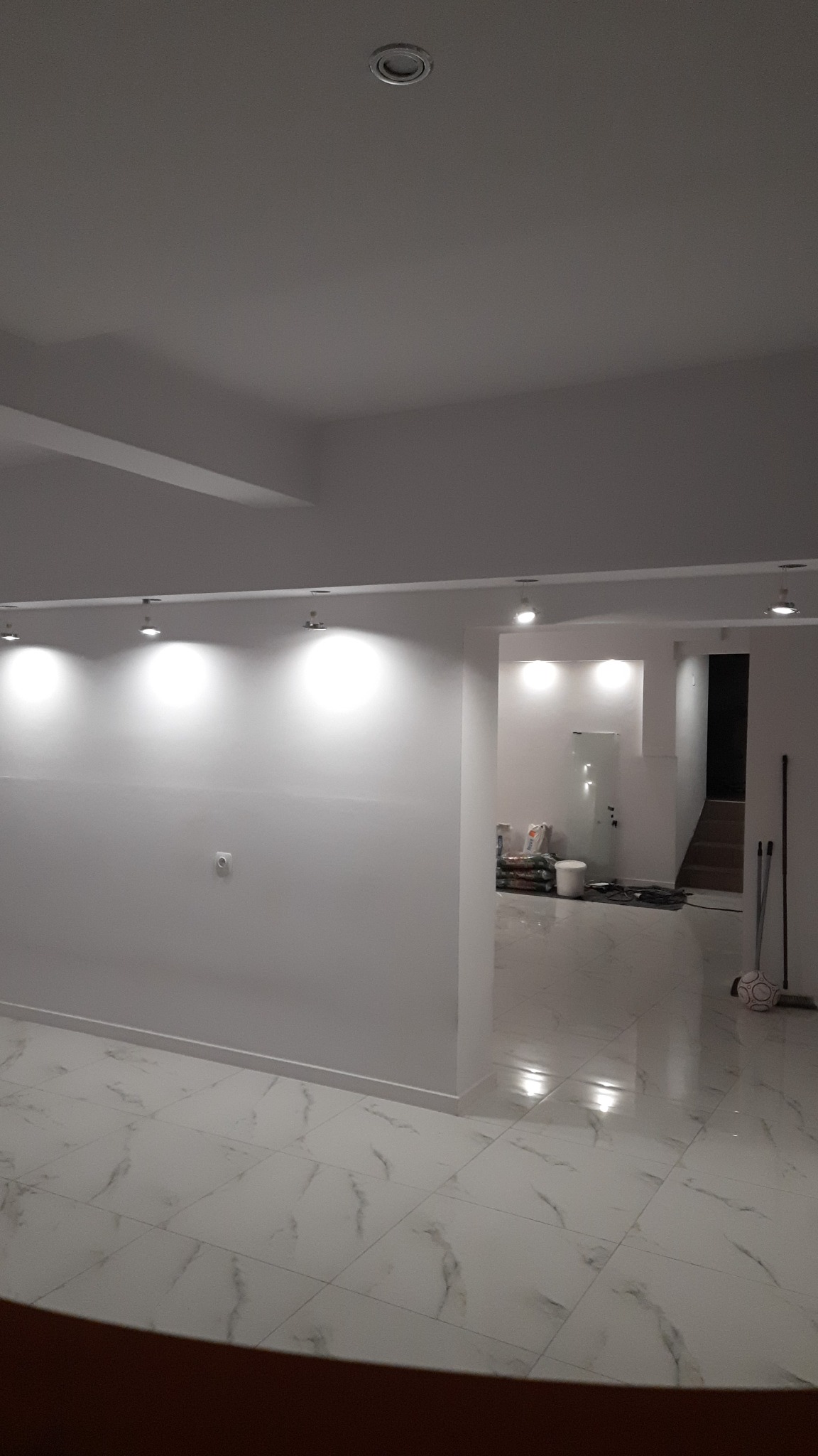 Białe, świeżo pomalowane wnętrze z marmurową podłogą i oświetleniem punktowym. Widoczne worki z materiałami budowlanymi w tle, sugerujące etap wykończeniowy.
