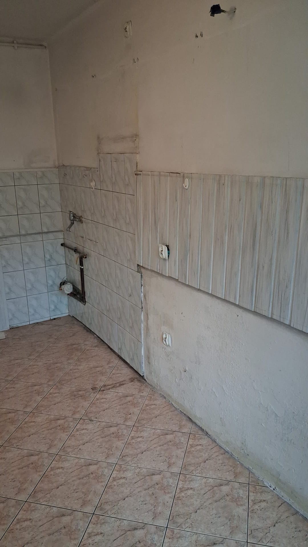 Wnętrze w trakcie prac przygotowawczych: częściowo położone płytki, fragment ściany z boazerią, widoczne instalacje. Podłoga wyłożona brązowymi płytkami.