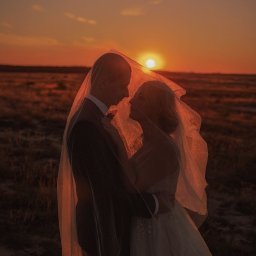 SASHA ART BEAUTY Oleksandra Melnychenko - Silueta panny młodej i pana młodego pod welonem na tle zachodzącego słońca. Romantyczny kadr z sesji ślubnej w plenerze, ciepłe barwy.