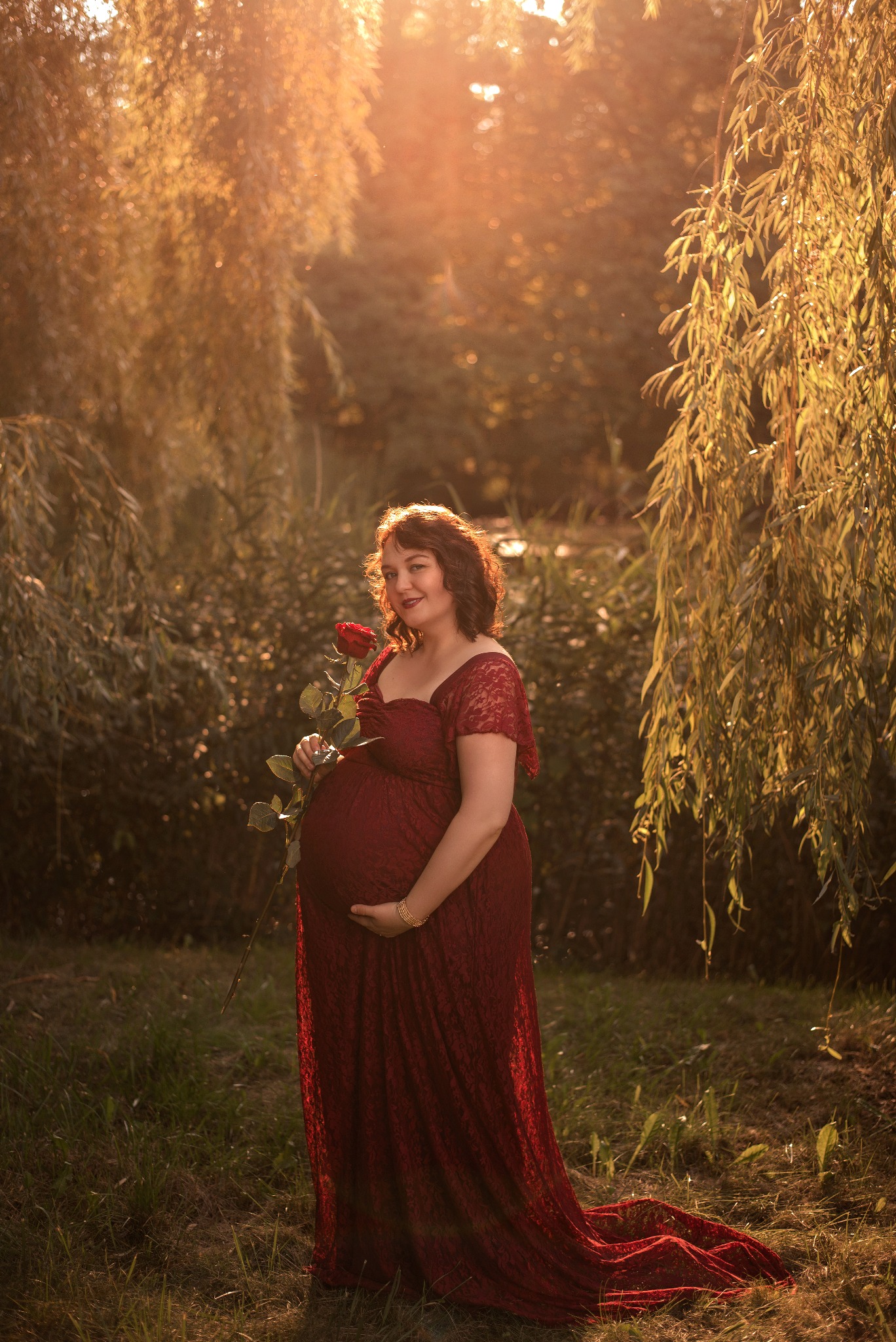 Kobieta w czerwonej sukni ciążowej, trzymająca czerwoną różę na tle drzew w promieniach zachodzącego słońca. Romantyczna sesja w plenerze.