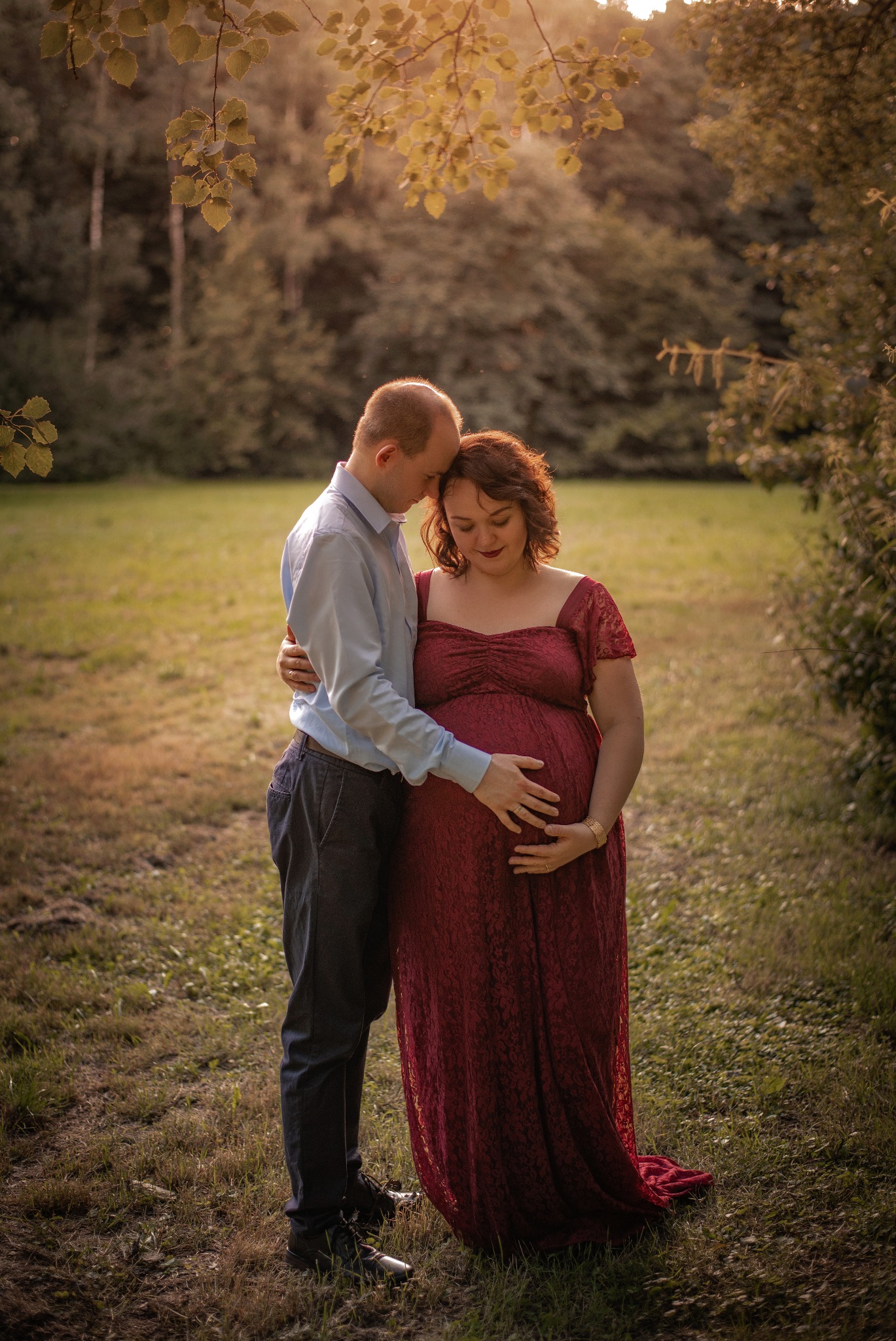 Ujęcie zakochanej pary na sesji ciążowej: kobieta w bordowej sukni, mężczyzna obejmuje ją, dłońmi dotykają brzucha na tle zieleni i ciepłego światła.