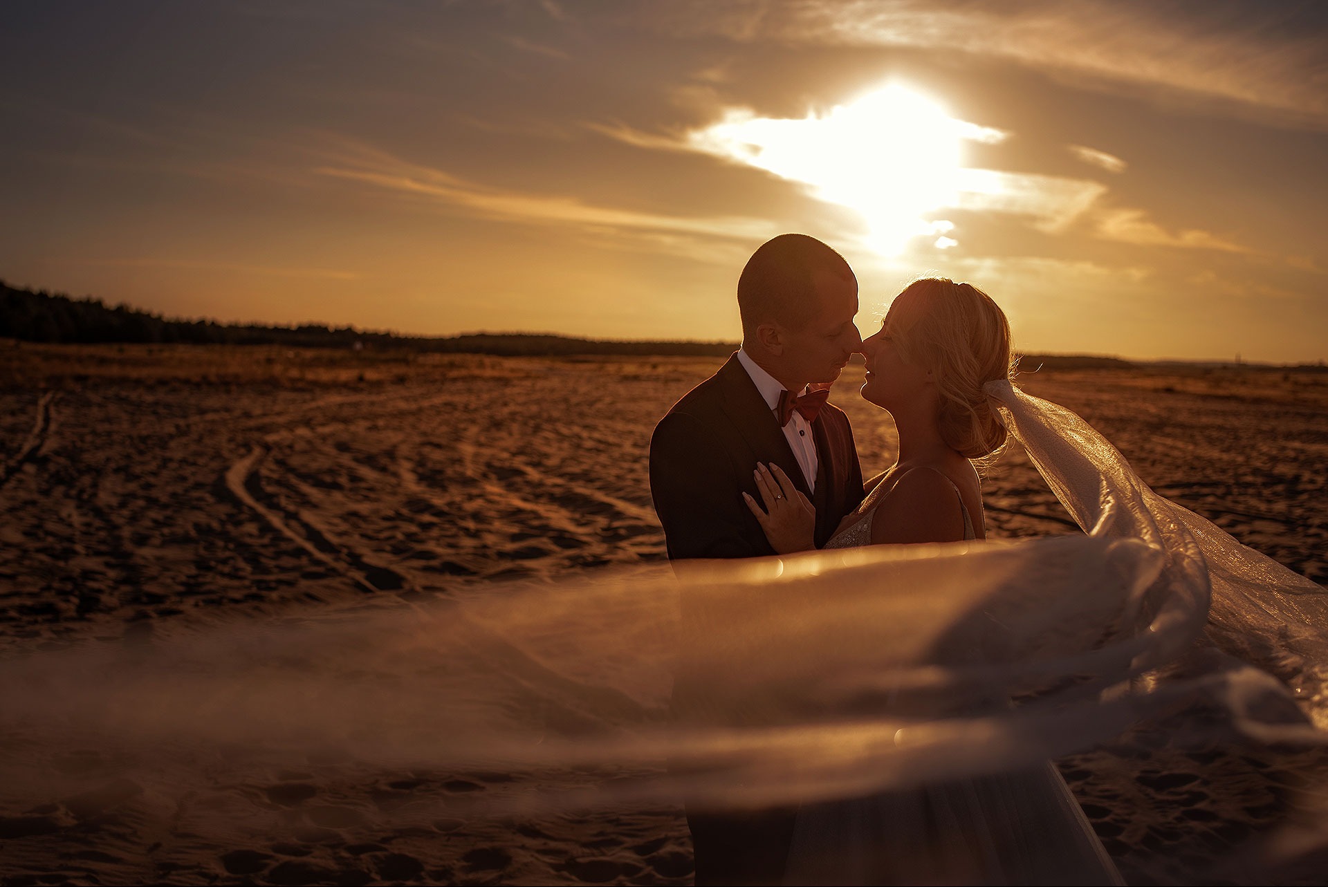 Para młoda w objęciach na tle zachodzącego słońca, welon delikatnie powiewa na wietrze. Ujęcie z dołu, podkreślające intymność chwili.