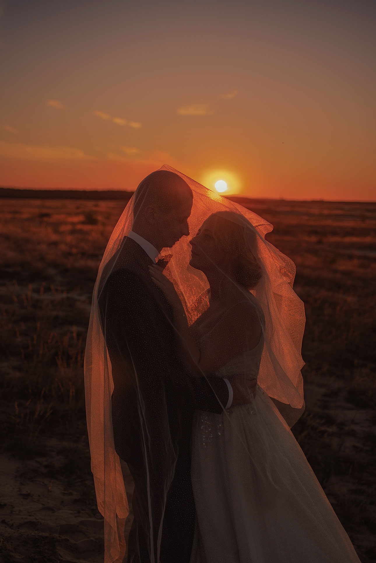 Silueta panny młodej i pana młodego pod welonem na tle zachodzącego słońca. Romantyczny kadr z sesji ślubnej w plenerze, ciepłe barwy.