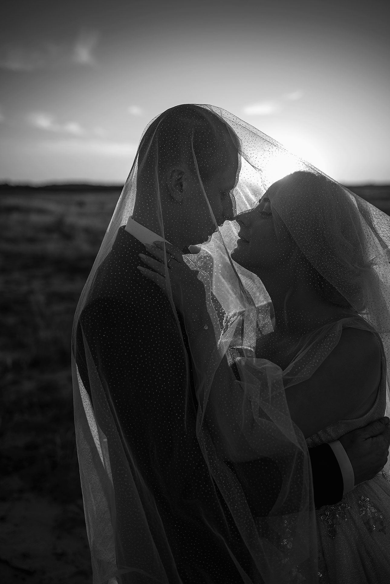 Czarno-białe zbliżenie pary młodej pod welonem na tle zachodzącego słońca. Pan młody w ciemnym garniturze, panna młoda z delikatnym makijażem. Romantyczna atmosfera.