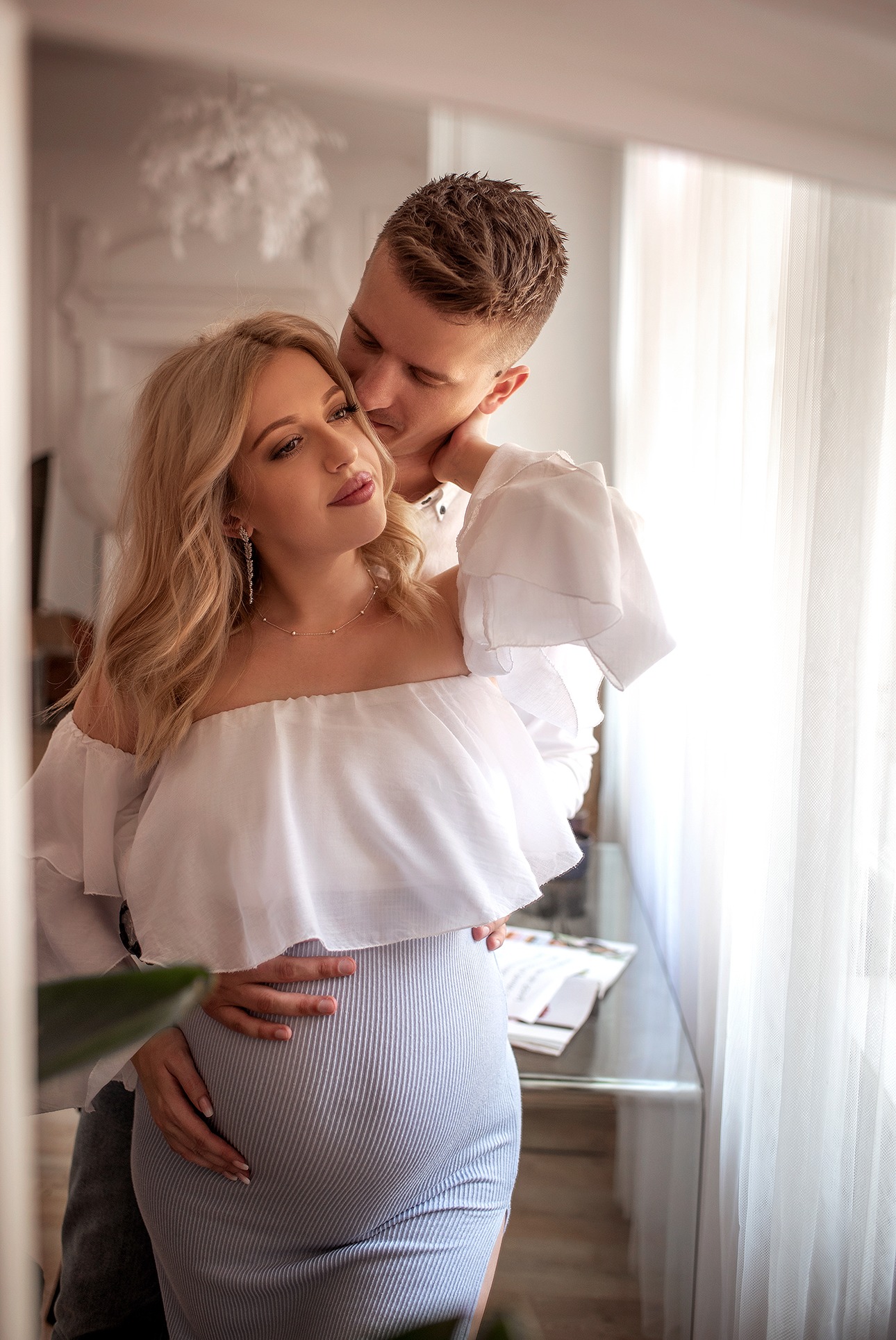 Ujęcie zakochanej pary: przyszła mama w błękitnej sukience i białej bluzce, czule obejmowana przez partnera w jasnym wnętrzu, delikatne światło.