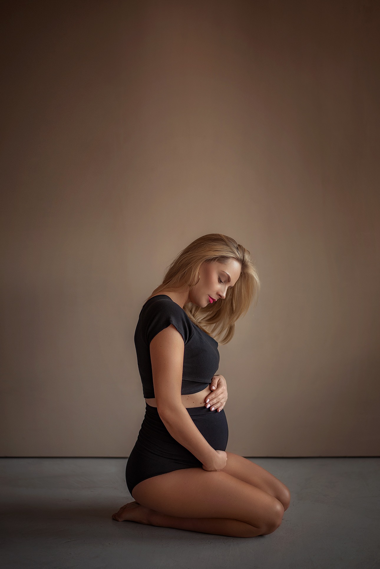 Kobieta w czarnym stroju, w pozycji klęczącej, delikatnie obejmuje ciążowy brzuszek na tle beżowego studia. Subtelne oświetlenie podkreśla naturalność i intymność.