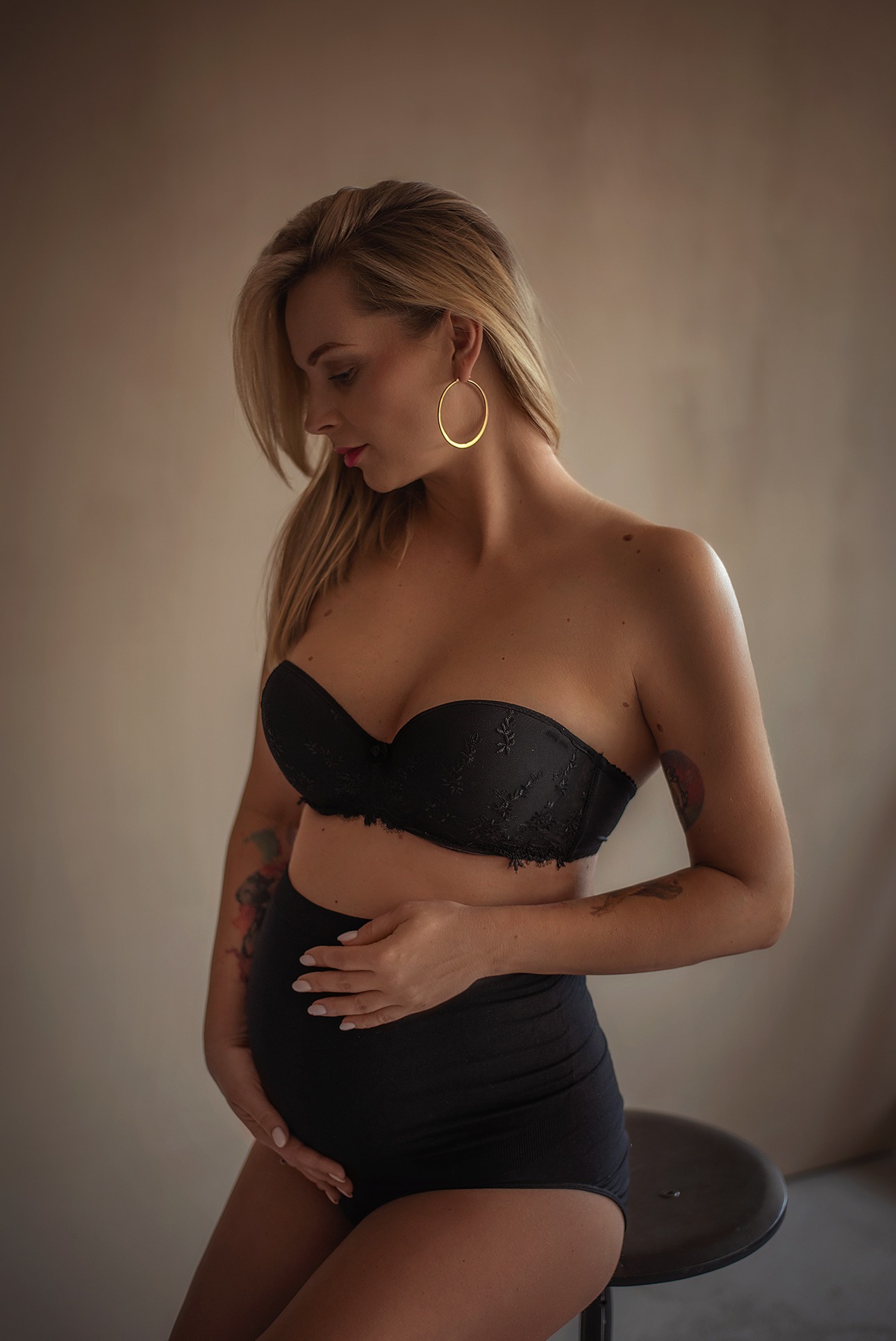 Kobieca fotografia ciążowa: blondynka w czarnej bieliźnie, z rękami na brzuchu, siedzi na stołku na neutralnym tle. Duże, złote kolczyki koła dodają blasku.