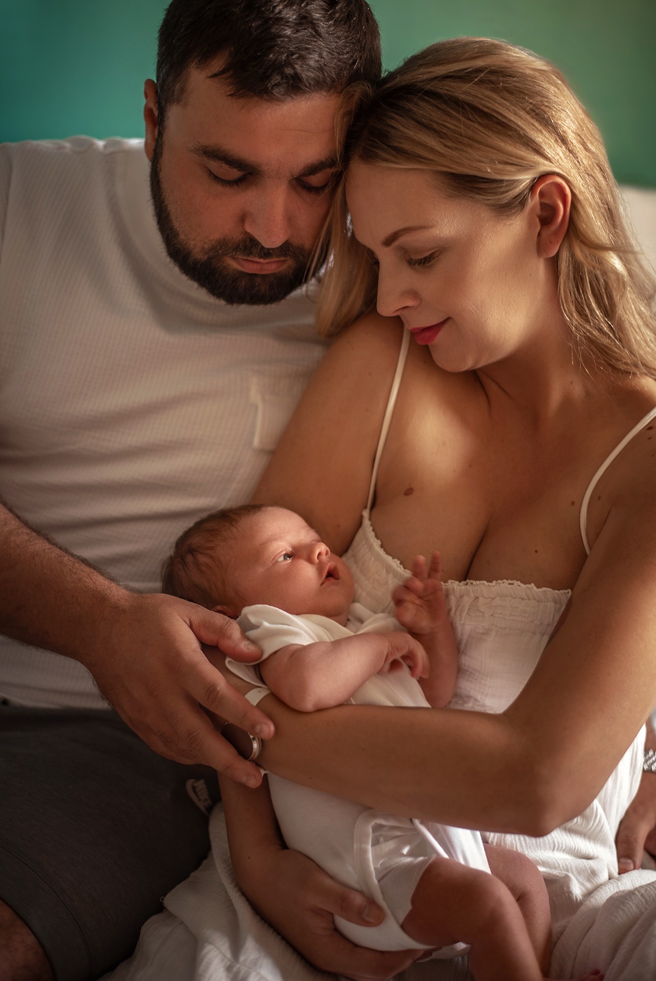 Rodzinne ujęcie: mama z blond włosami i tata z brodą tulą noworodka w białym ubranku, patrząc z miłością. Delikatne światło, ciepłe kolory, intymna atmosfera.