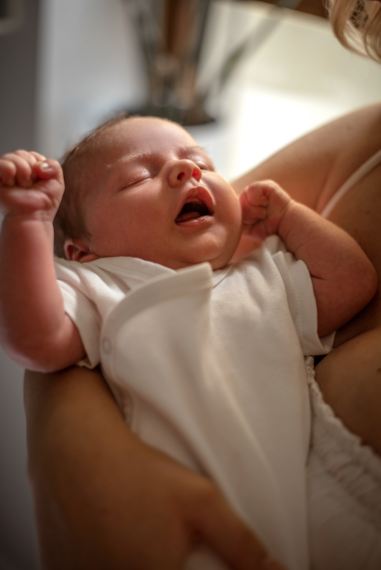 Noworodek w białym ubranku, z zaciśniętymi piąstkami, leży na ramionach matki. Delikatne światło podkreśla detale twarzy dziecka.