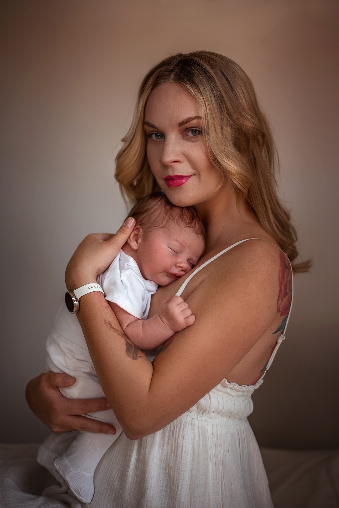 Blondwłosa mama w białej sukience tuli śpiącego noworodka w białym ubranku na tle beżowej ściany. Widoczny tatuaż na ramieniu kobiety i zegarek na jej nadgarstku.