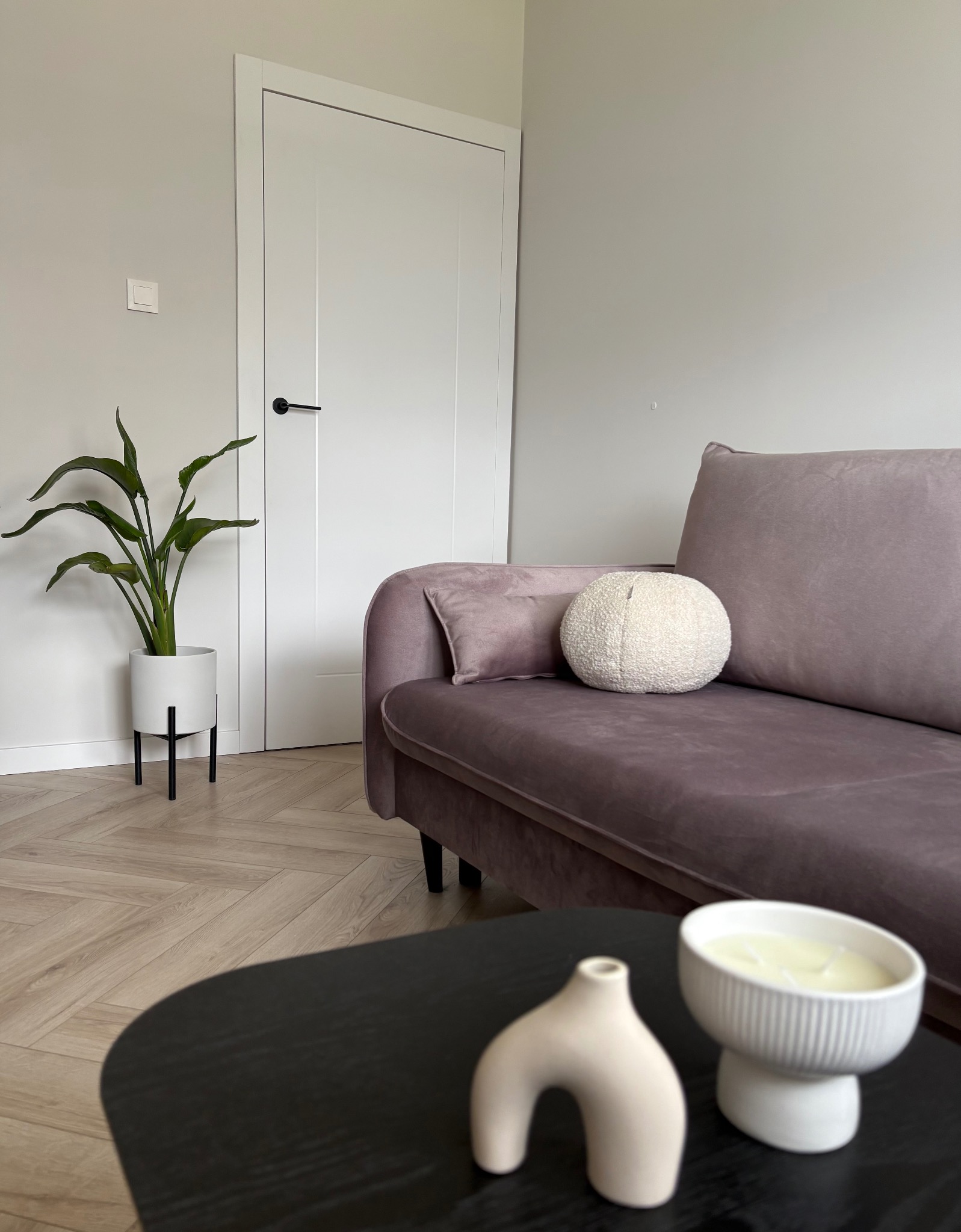 Białe, minimalistyczne drzwi wewnętrzne z czarną klamką w nowoczesnym salonie z fioletową sofą i drewnianą podłogą w jodełkę.