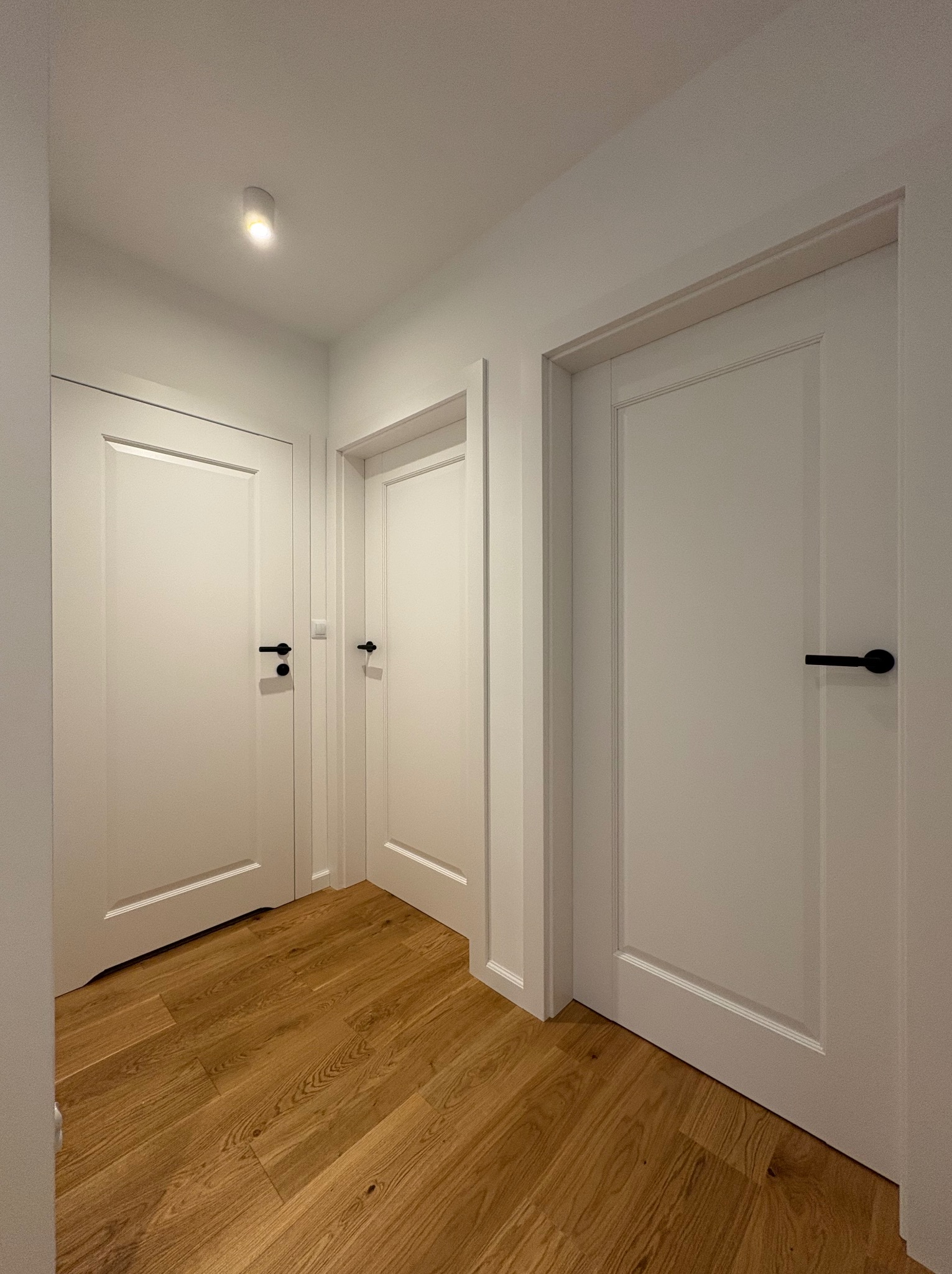 Białe drzwi wewnętrzne z czarnymi klamkami w jasnym korytarzu z drewnianą podłogą. Minimalistyczny design, równomierne oświetlenie, widok na trzy skrzydła drzwiowe.