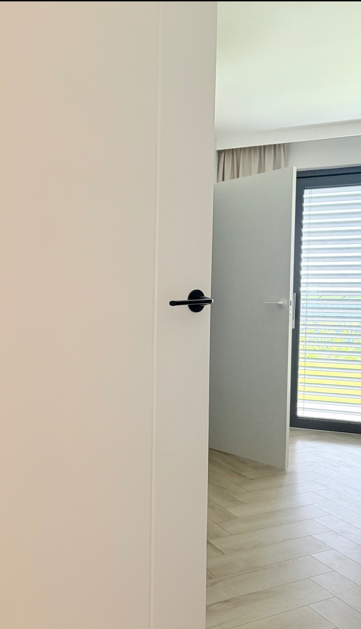 Otwarta, biała płaszczyznowa konstrukcja z czarną klamką w nowoczesnym wnętrzu. W tle widoczne okno z żaluzjami i parkiet ułożony w jodełkę.