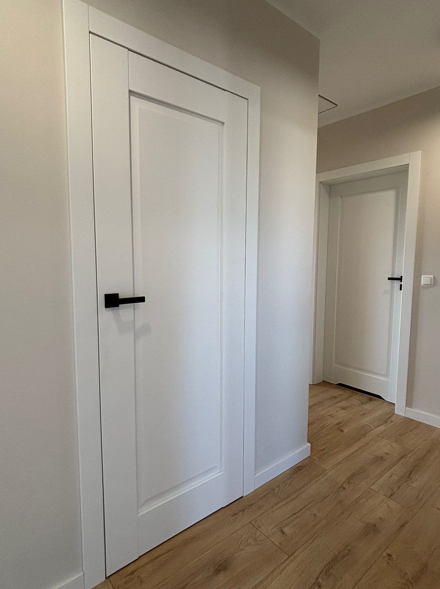 Białe drzwi wewnętrzne z czarną klamką w minimalistycznym wnętrzu. Widoczne drewniane panele podłogowe i fragment drugich drzwi w tle.