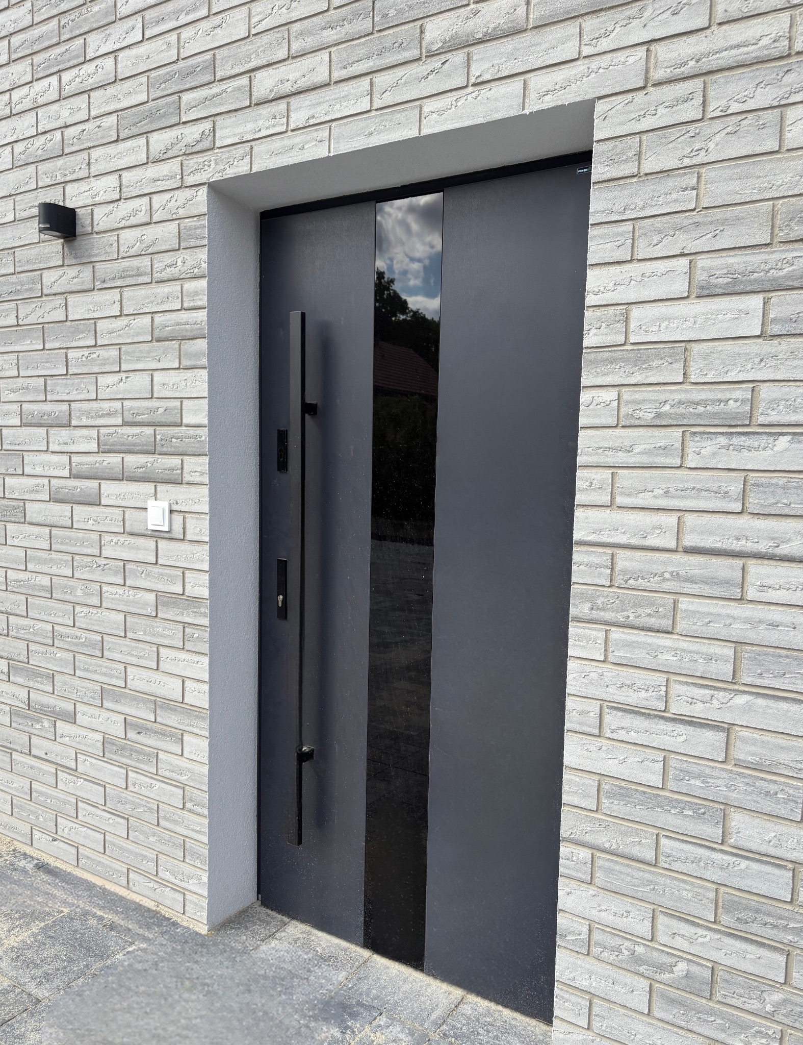Nowoczesne, grafitowe drzwi zewnętrzne z długim uchwytem, wkomponowane w elewację z białej cegły. Centralnie umieszczony panel szklany odbija otoczenie.