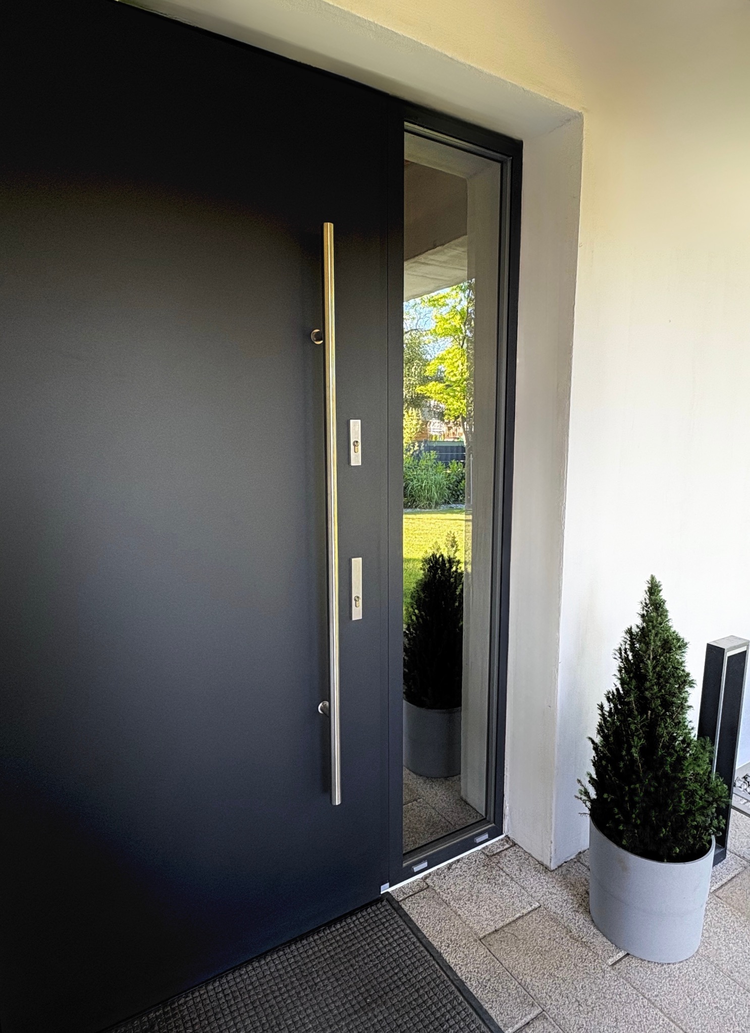 Nowoczesne, czarne drzwi zewnętrzne z długim, srebrnym uchwytem i szybą boczną, obok donica z iglakiem, widok z zewnątrz.