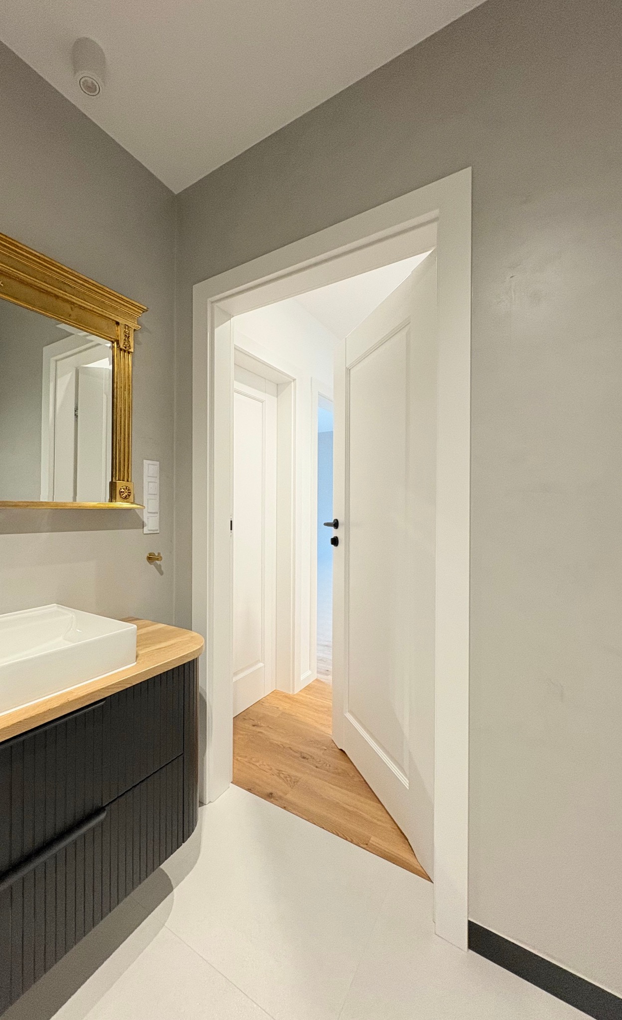 Otwarta biała płycinowa  wewnętrzna z czarną klamką. W tle widoczna podłoga z desek. Obok umywalka z lustrem w złotej ramie. Szare ściany.