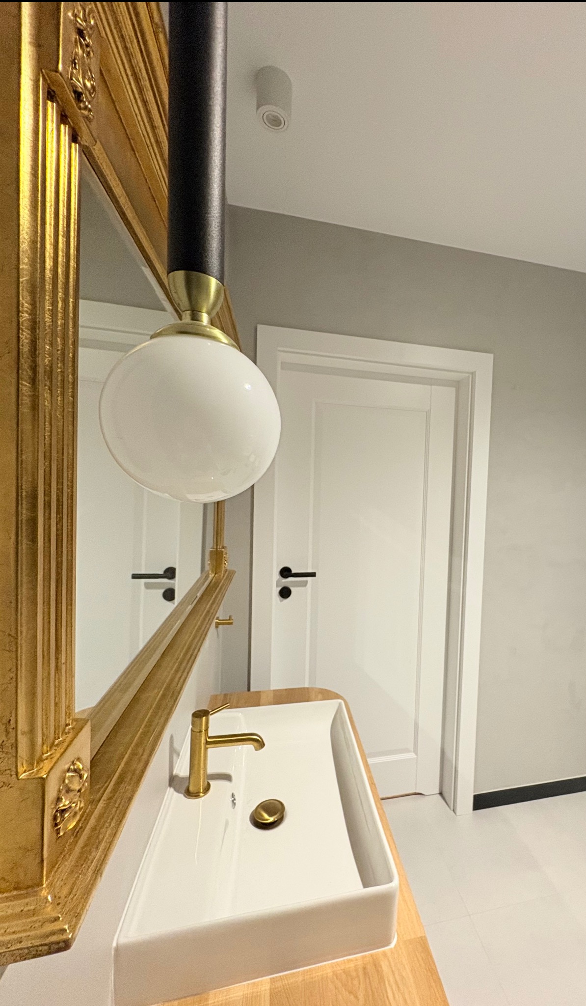 Nowoczesna łazienka z białą umywalką na drewnianym blacie, złotą armaturą i lustrem w bogato zdobionej, złotej ramie. W tle białe drzwi z czarną klamką.