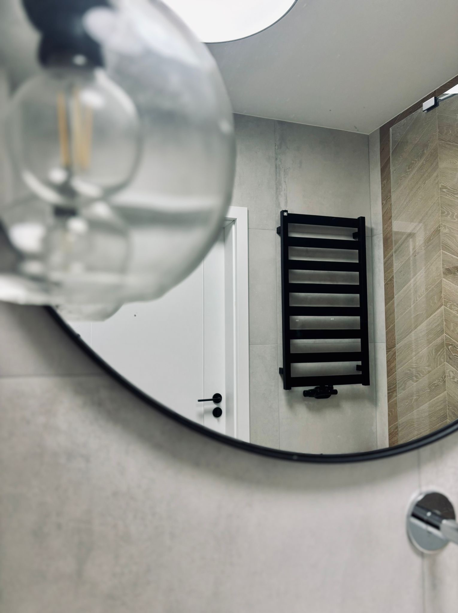 Lampa i białe drzwi z czarną klamką, widziane w odbiciu okrągłego lustra w nowoczesnej łazience z czarnym grzejnikiem drabinkowym.