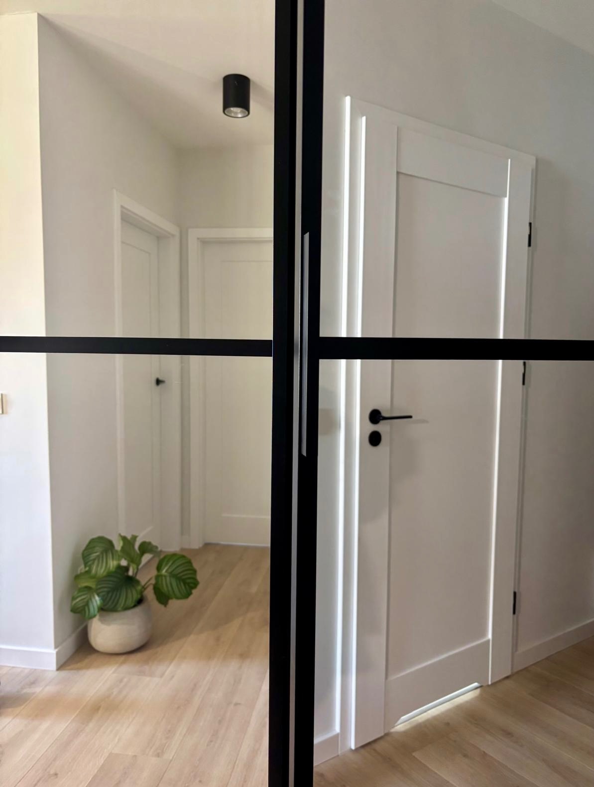 Nowoczesne drzwi szklane z czarną ramą, dzielące przestrzeń. W tle widoczny korytarz z białymi drzwiami i rośliną doniczkową. Jasne wnętrze, parkiet w jodełkę.