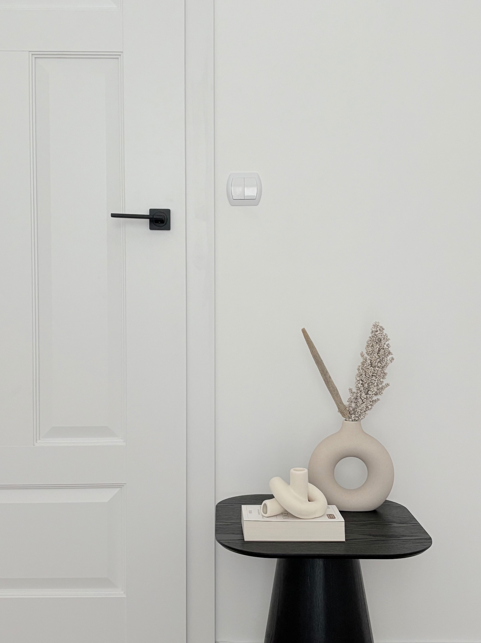 Białe drzwi wewnętrzne z czarną klamką, obok stolik z dekoracjami w minimalistycznym stylu. Widoczny włącznik światła na ścianie.