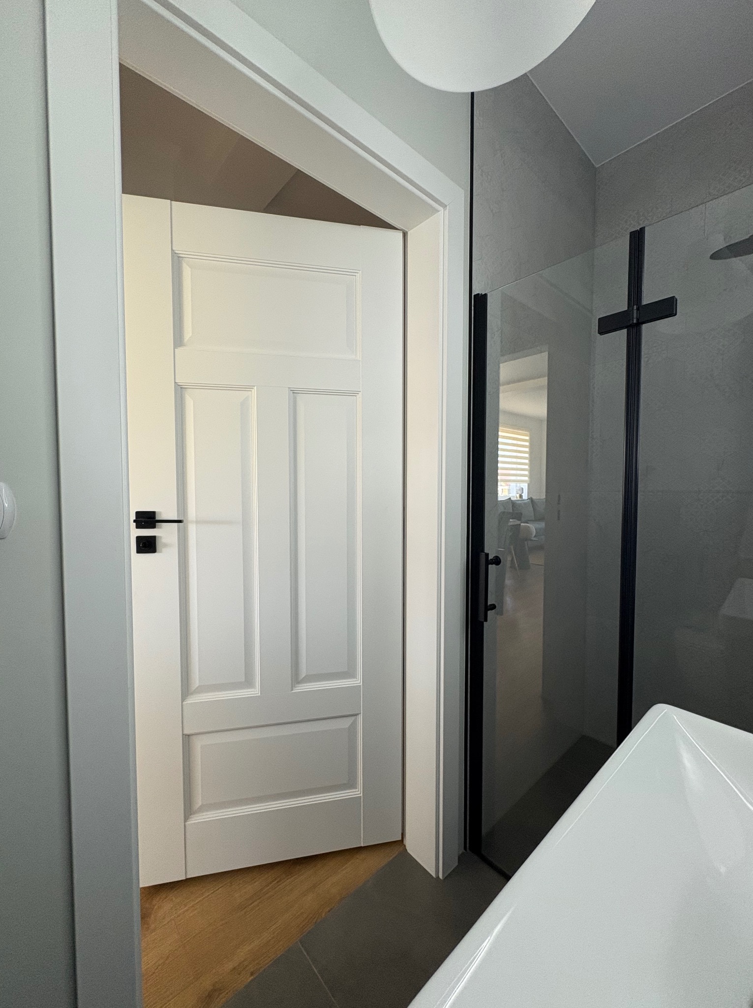 Białe, częściowo otwarte drzwi z czarną klamką, widoczne z wnętrza łazienki z kabiną prysznicową i umywalką. Podłoga w dwóch odcieniach szarości i brązu.
