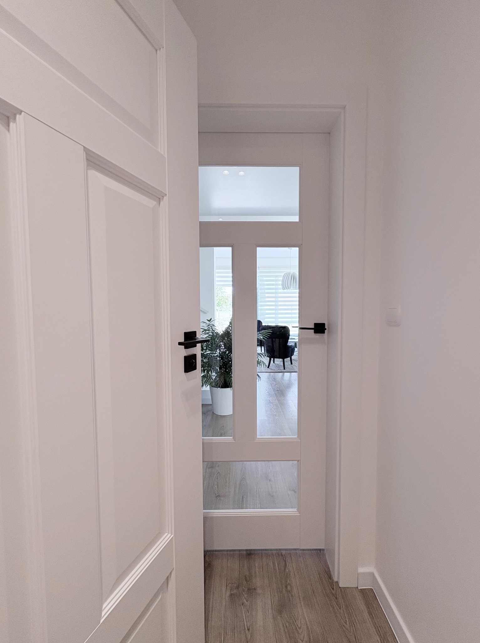 Białe drzwi wewnętrzne z przeszkleniami i czarną klamką, widok z korytarza na salon. Minimalistyczny design, jasne wnętrze. Parkiet w odcieniach szarości.