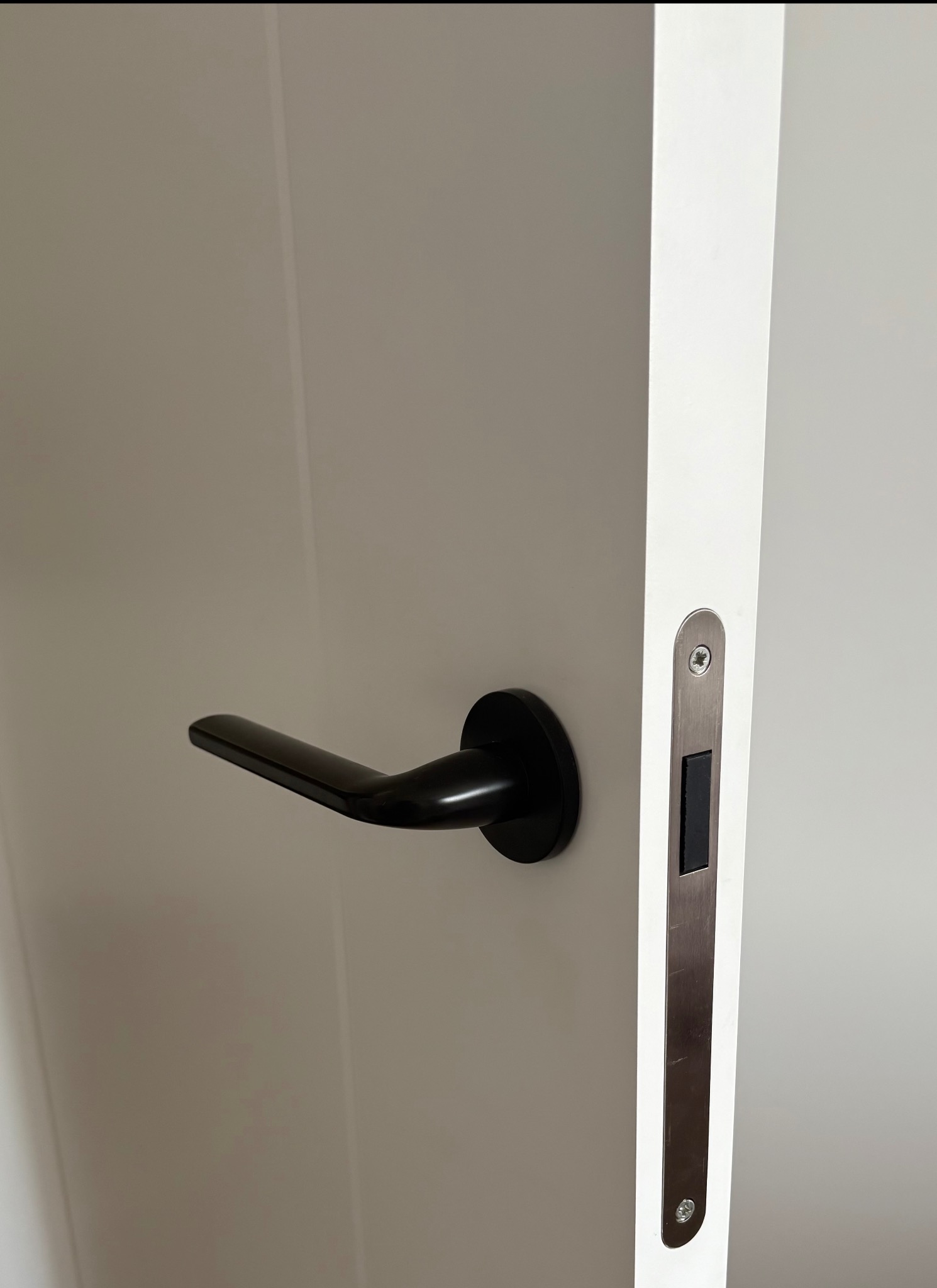 Białe skrzydło drzwiowe z czarną klamką i metalowym zamkiem w srebrnej obudowie. Minimalistyczny design, gładka powierzchnia, nowoczesne wykończenie.